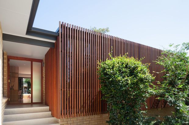 Exterior view of a contemporary residential entry with a covered walkway, broad concrete steps, warm timber-framed glazing, and a prominent vertical timber batten privacy screen above a low brick wall. Mature shrubs partially screen the facade, while a dark projecting roof edge and exposed structural beams define the entrance composition.