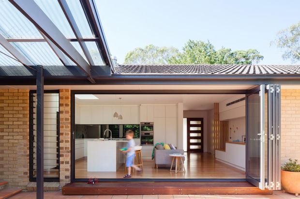 Bright view into a contemporary single-storey brick home through fully opened folding glazed doors. The image shows an open-plan kitchen and living area, with a white island, integrated white cabinetry, timber flooring, a grey sofa, and a covered outdoor transition zone with timber decking. A large translucent-roofed pergola or verandah structure frames the left side, while the tiled roof and exposed brick facade are visible around the opening.