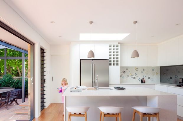 Bright contemporary kitchen with a large white island and three stools, stainless-steel double-door refrigerator, white cabinetry, patterned tile splashback, pendant lights, skylight, and timber flooring. A glazed opening connects to an outdoor patio, and a child is visible near the kitchen.