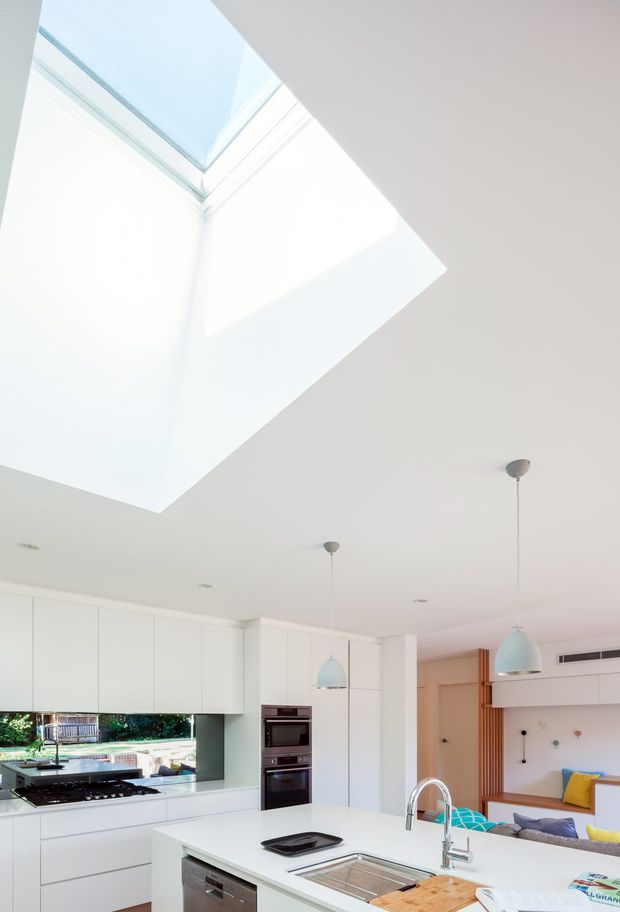 Bright contemporary open-plan kitchen viewed beneath a large rectangular skylight. The space features extensive white flat-panel cabinetry, a white island with integrated sink and dishwasher, stainless-steel tapware and appliances, pale blue pendant lights, and a connected living area with built-in joinery. Strong natural light creates an airy, clean interior.