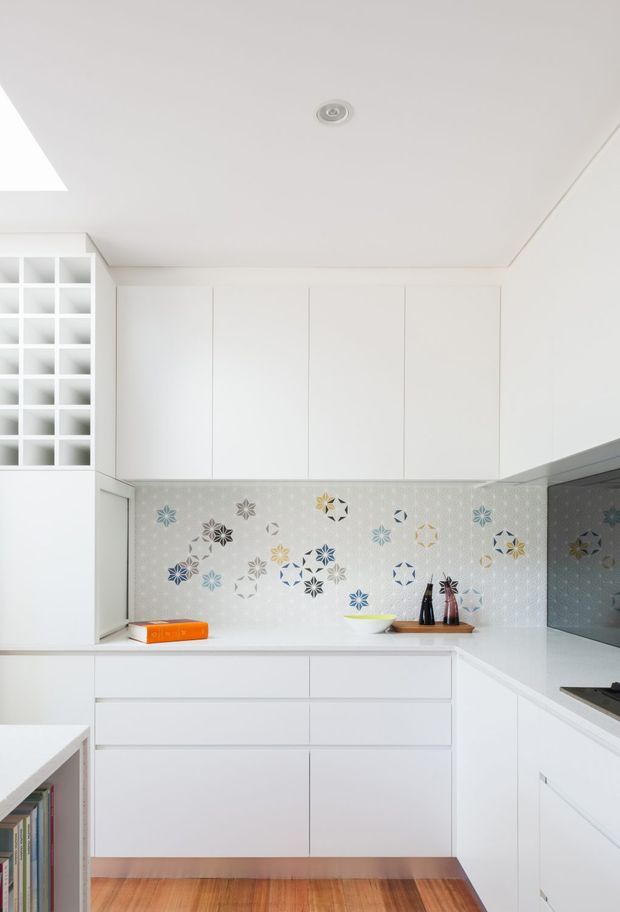 Bright, compact contemporary white kitchen with flat-panel cabinetry, white benchtops, and a highly decorative patterned splashback featuring geometric floral motifs. A white open-grid partition is visible on the left, with timber flooring and a dark reflective window or appliance surface on the right. The space is clean and predominantly white with small colourful splashback accents.