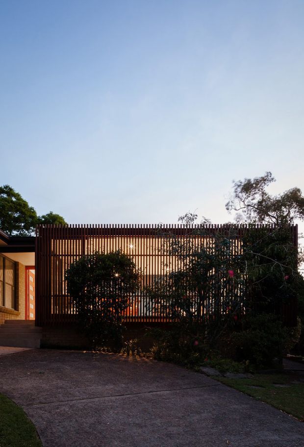 Exterior view of a contemporary single-storey residence at dusk, showing a broad driveway, a recessed entry, warm timber-toned walls, and a prominent vertical timber screening facade. Dense mature planting partially conceals the building, while warm interior lighting glows through the slatted screen.