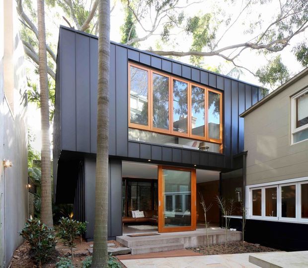 Exterior view of a contemporary multi-level residential extension with dark vertical metal cladding, warm timber-framed glazing, and a recessed ground-floor entrance. The building is integrated among mature trees, with a paved entry path and a neighbouring light-coloured dwelling visible at right.