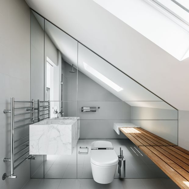A compact contemporary bathroom beneath a sloping roof, featuring grey large-format tiled walls and floor, extensive frameless mirror panels, a wall-hung white toilet, floating marble-effect vanity, chrome heated towel rail, and a long timber bench or shelf along the angled wall. A skylight brings in natural light.