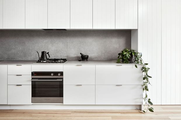 A clean, modern galley-style kitchen with white flat-panel cabinetry extending across the wall, integrated lower drawers and cupboards, a built-in stainless-steel oven, and a gas cooktop. A continuous grey stone-look backsplash and benchtop create a restrained contrast, while pale timber flooring adds warmth. A trailing green plant and dark kettle provide small accents.