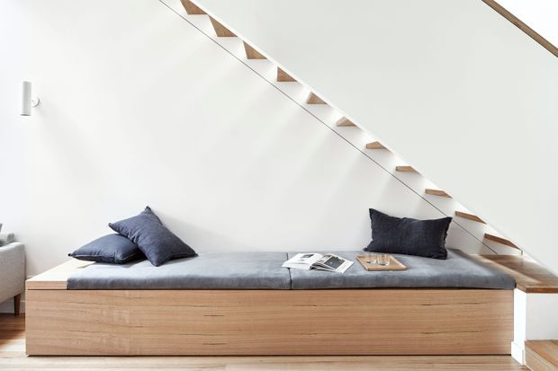 Bright minimalist interior beneath a floating open-riser staircase. A long built-in timber bench or daybed with pale grey cushioning and dark blue cushions sits against a white wall, with a small reading lamp at left and books placed on the seat. The staircase has slim timber treads, a white stringer or side structure, and a simple linear handrail.
