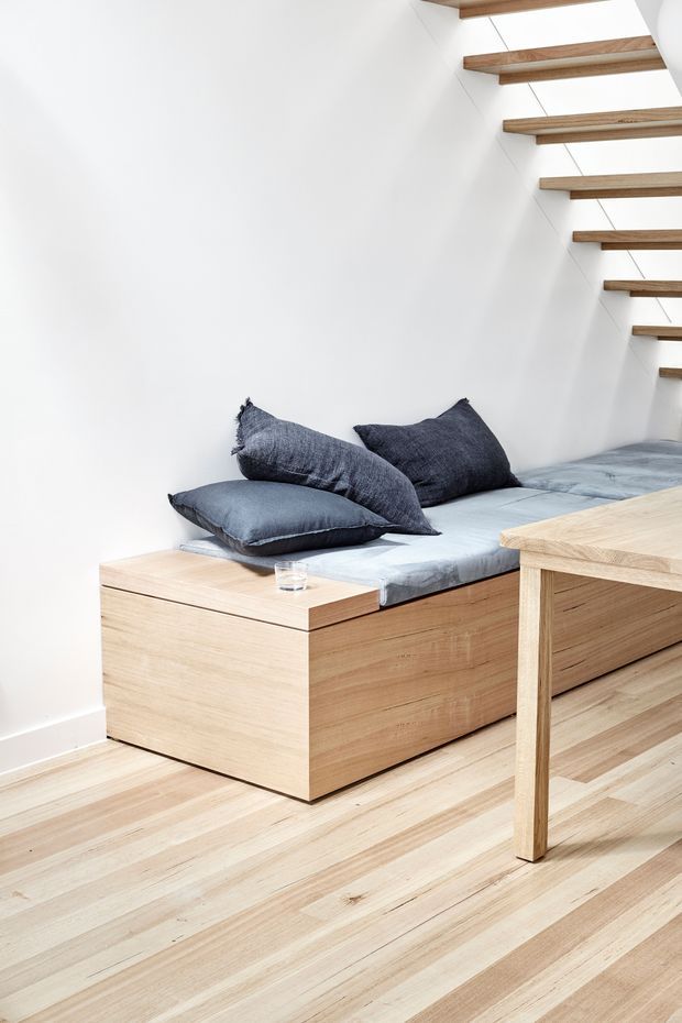 Bright minimalist interior featuring a built-in timber daybed with blue-grey cushions beneath an open-riser timber staircase. Pale timber flooring and a simple light-coloured wall create a clean, compact multi-purpose seating area, with part of a timber table visible in the foreground.