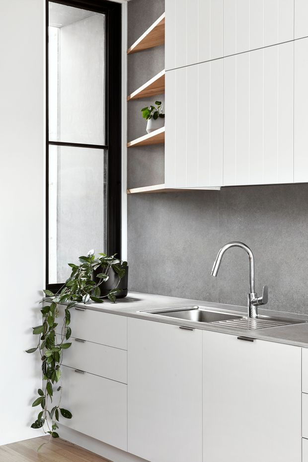 A compact contemporary kitchen with white flat-panel cabinetry, a grey stone-look backsplash, stainless-steel sink and drainer, and a tall chrome gooseneck tap. Open timber shelves with dark backing add warmth beside a narrow black-framed window, while trailing indoor plants soften the minimalist scheme. Light timber flooring is visible at the base.