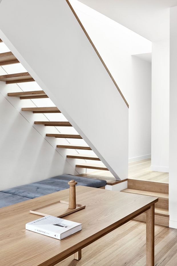 Bright minimalist interior showing an open-riser timber staircase with a white sculptural stair enclosure, pale timber flooring, and a large simple timber table in the foreground. A built-in blue upholstered seating area sits beneath the stairs, with strong natural light entering from above and through the stair void.