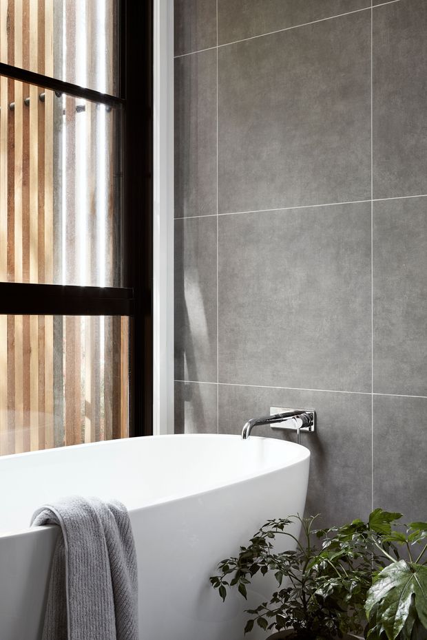 A close-up contemporary bathroom scene featuring a freestanding white bathtub, wall-mounted chrome tapware, large grey wall tiles, and a dark-framed window with vertical timber screening beyond. A leafy indoor plant and grey towel add softness to the otherwise clean, restrained space.
