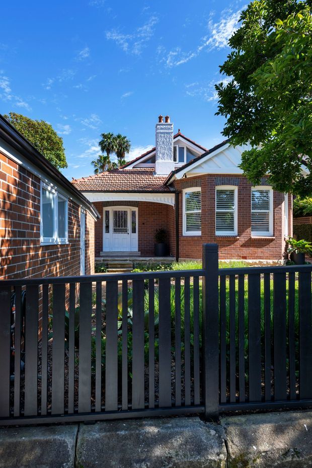 Bright exterior view of a traditional red-brick residential cottage or bungalow behind a black vertical-slat front gate. The house has a terracotta tiled pitched roof, a prominent rendered chimney with twin pots, white-framed windows with horizontal blinds, a covered entry porch, and a small landscaped front garden. Mature trees and palms surround the property under a clear blue sky.