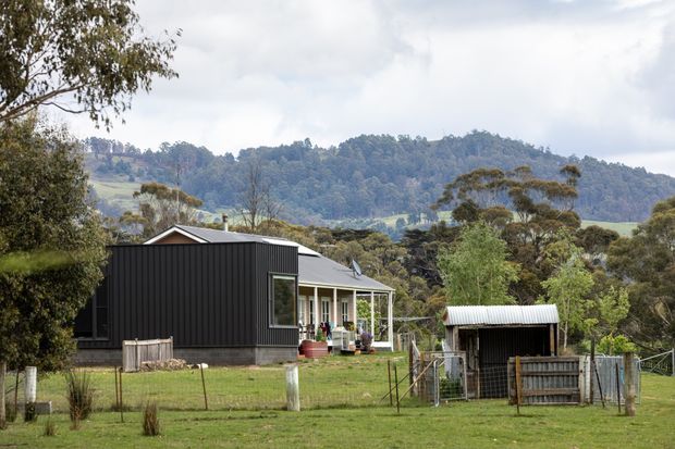 Exterior view of a rural contemporary home with a dark vertically clad extension, light-colored roof, and a covered veranda. The property sits in a grassy fenced setting with a small corrugated-metal outbuilding, surrounded by mature trees and rolling wooded hills.