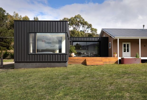 A low, single-storey contemporary residential extension or compact home composed of contrasting black vertical metal-clad volumes and a lighter gabled section. Large glazed openings connect the central living area to a raised timber deck, while the front lawn and surrounding mature trees create a rural or suburban setting.