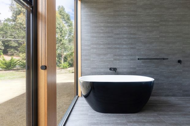 A contemporary bathroom featuring a freestanding black-and-white oval bathtub against a large-format grey tiled wall. A slim black wall-mounted tap and bath filler are paired with a timber-framed full-height window, providing views to a landscaped outdoor setting. Grey tiled flooring and restrained black fixtures create a refined, minimalist atmosphere.