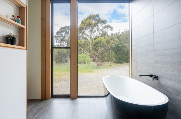 A contemporary bathroom featuring a freestanding white oval bathtub with a dark exterior, positioned beside full-height timber-framed glazing overlooking a grassy rural landscape. The room has large grey wall and floor tiles, a minimalist black wall-mounted tap, and built-in timber shelving and cabinetry.