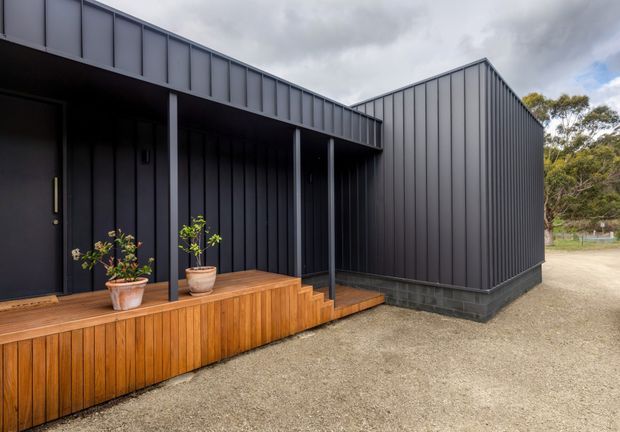 Exterior view of a contemporary dark-clad building with strong vertical metal wall panels, a low sloping roofline, and a covered entrance verandah. The entry is approached by a warm timber deck and steps with potted plants, while a gravel forecourt surrounds the structure. Native vegetation is visible in the background beneath an overcast sky.