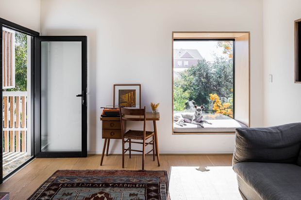 Bright, compact contemporary living room or entry sitting area with pale walls and light timber flooring. A black-framed glazed door opens to an exterior balcony or porch, while a large timber-framed window overlooks greenery. The room includes a dark upholstered sofa, a small mid-century-style timber writing desk and chair, framed artwork, and a patterned rug. Natural daylight creates a warm, inviting atmosphere.