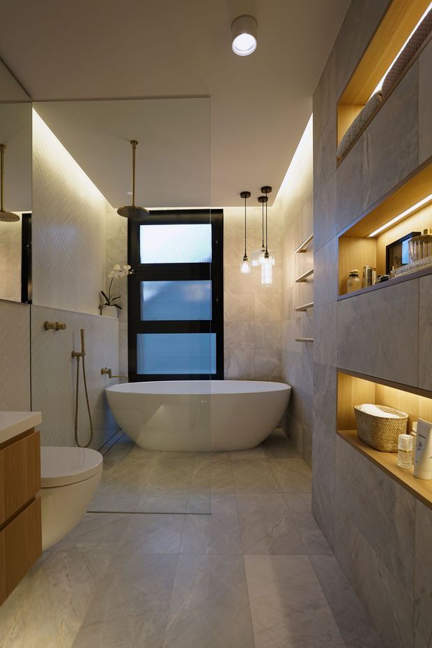 A contemporary bathroom with a freestanding oval bathtub beneath a frosted black-framed window, a wall-mounted shower, toilet, and integrated illuminated display niches. The space uses large-format pale stone-look tiles, warm timber-toned joinery, recessed shelving, pendant lights, and soft indirect lighting.