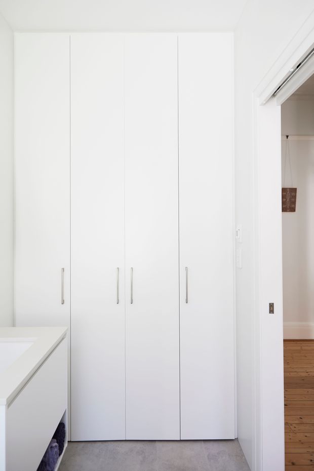 A bright, minimalist built-in storage wall with tall white full-height cabinet doors and slim vertical metal handles. A white benchtop or island edge is visible at lower left, while an open doorway reveals a timber-floored adjacent room. The cabinetry is set against white walls with a grey smooth floor finish.