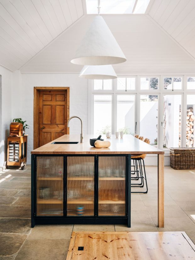 Bright, airy kitchen centered on a large timber-topped island with black-framed ribbed glass cabinetry, integrated sink and seating. The room features white painted timber wall and ceiling panelling, a pitched vaulted ceiling with skylight, oversized white pendant lights, a timber entry door, expansive white-framed windows and pale stone or tile flooring. The overall character is warm, light and refined with a blend of traditional architectural detailing and contemporary joinery.