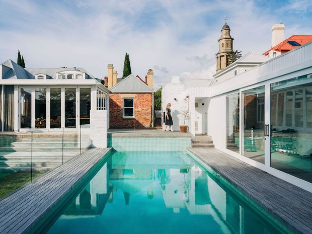 A bright architectural courtyard centered on a narrow turquoise swimming pool, surrounded by white contemporary glazed pavilions and timber decking. A historic brick building and ornate clock tower are visible beyond the courtyard, creating a contrast between modern additions and heritage architecture. The scene has abundant glazing, crisp white surfaces, reflective water, and a sunny open-air atmosphere.