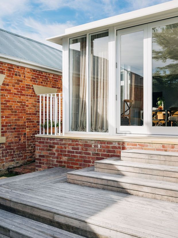 Exterior view of a brick-clad house extension opening onto a stepped timber deck. White-framed floor-to-ceiling glazing and a glazed door connect the interior to the outdoor platform, with a small white balustrade beside the steps. The scene is bright and contemporary, combining red brick, pale weathered timber decking, and white trim.