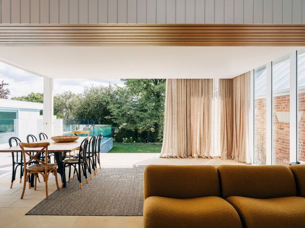 A bright, open-plan living and dining space connected to a landscaped outdoor area through extensive glazing. The room features a warm mustard-brown curved sofa, timber dining table and chairs, black dining chairs, full-height beige curtains, a textured neutral rug, pale flooring, and a distinctive timber-lined ceiling bulkhead with linear detailing. Brickwork is visible through the windows, while the interior has a refined contemporary character.