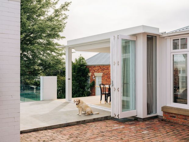 Exterior view of a contemporary white-framed glazed extension opening onto a paved brick terrace. The image shows a flat-roofed covered outdoor area, large folding glass doors, white rendered walls, and an adjacent red-brick building and paving. A dog and surrounding vegetation are visible but are not part of the architectural focus.