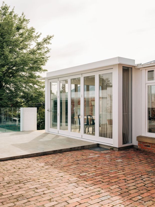 Exterior view of a bright contemporary home extension opening onto a brick-paved terrace. The glazed room features large white-framed folding or sliding doors, pale vertical cladding, a flat roof, and a simple concrete patio beside a glass pool or balustrade area. Mature trees and an adjoining brick-and-painted exterior are visible.