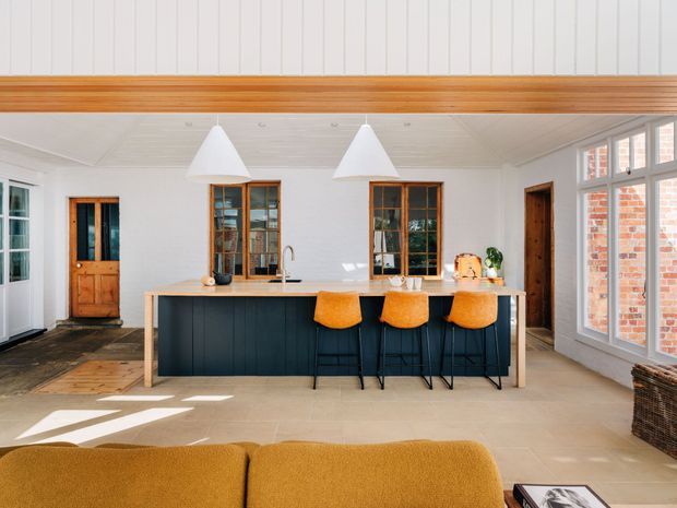Bright open-plan kitchen and living area with a large dark-panelled island, pale stone benchtop, four orange upholstered bar stools, white pendant lights, timber-framed windows and doors, white vertical wall panelling, and a timber ceiling beam. Large glazed windows reveal exposed brick outside, while the foreground includes part of a mustard sofa.