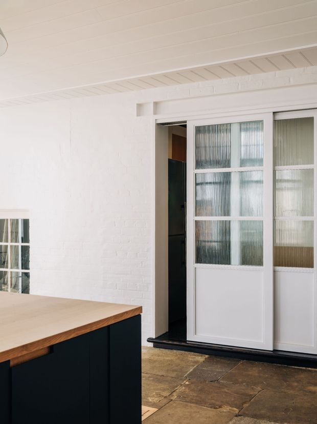 Bright interior view featuring a white-painted brick wall, white paneled ceiling, and a white-framed glazed partition or door with ribbed/frosted glass. A dark built-in island or counter with a light timber benchtop occupies the foreground, while dark stone or concrete flooring contrasts with the pale surfaces. The space has a clean, restrained contemporary character with subtle industrial detailing.