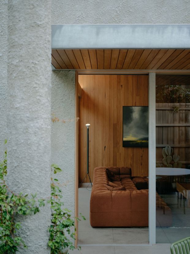 A close architectural view through a glazed opening into a warm, timber-lined living room. The exterior wall has a heavily textured pale stucco finish with climbing greenery, while the entry and ceiling soffit are clad in vertical and slatted timber. A large brown leather sectional, slender floor lamp and framed artwork are visible inside, with a concrete-toned floor and a glimpse of a courtyard fence beyond.