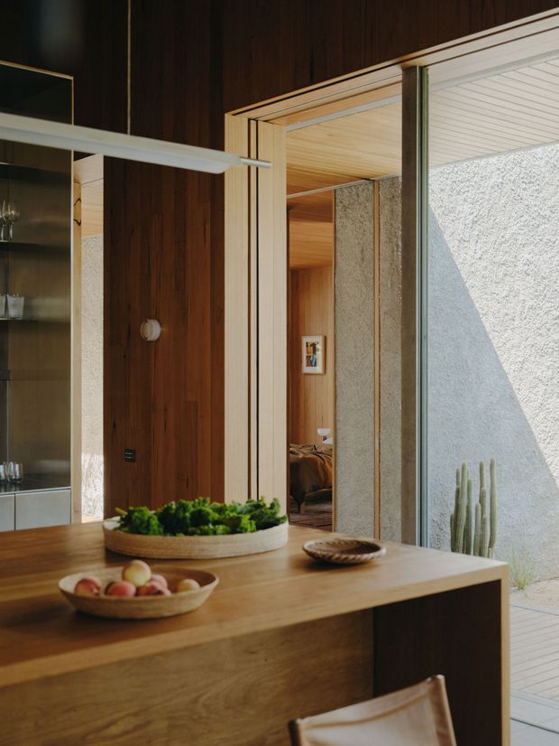Warm, wood-lined interior centered on a timber kitchen or dining island with a bowl of fruit and a planter. A large mirror reflects the room, while an open timber-framed passage leads toward a bedroom or living area. Floor-to-ceiling glazing opens to a bright textured courtyard with a cactus, and the ceiling features light timber boards. The overall composition is calm, natural, and strongly connected to the outdoors.