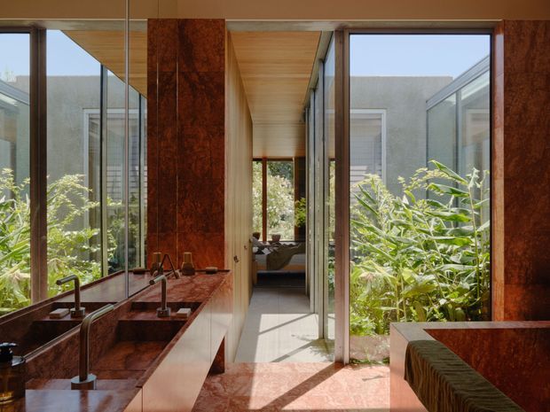 A refined contemporary bathroom with a long floating vanity, twin basins and polished reddish-brown stone surfaces. Full-height glazing and mirrored panels open the room visually to lush planting, while a timber-lined ceiling and corridor lead toward a bedroom or living space beyond. The space is bright, warm, and strongly connected to greenery.