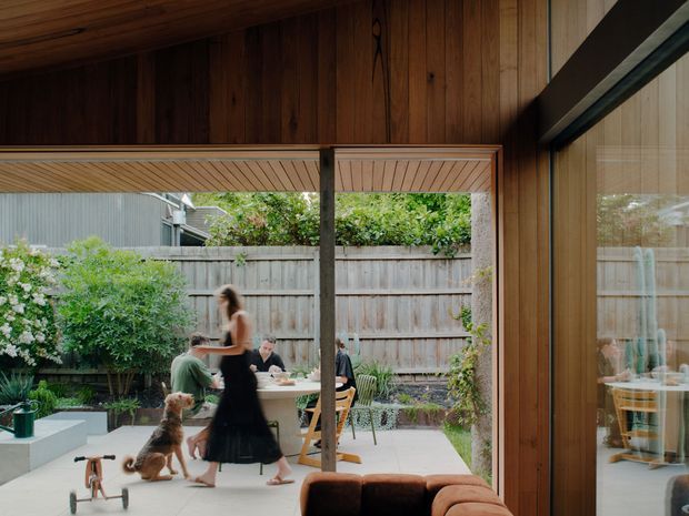 View from a timber-lined interior opening onto a landscaped courtyard dining area. The space features warm vertical and horizontal timber lining, large glazed sliding doors, a broad covered opening, concrete paving, outdoor dining furniture, planting, and a timber fence. The image presents a strong indoor-outdoor connection with people dining in the courtyard and reflections visible in the glazing.