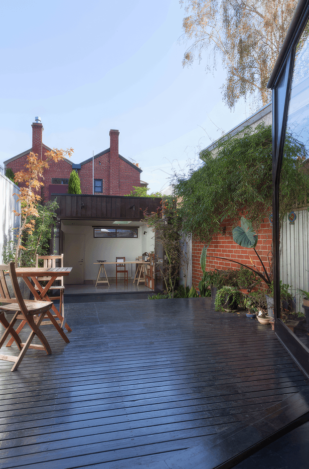 A sunlit urban courtyard and elevated timber deck viewed from beside full-height glazing. The dark stained deck extends toward a compact covered outdoor workspace or entertaining area, with a timber dining table and folding chairs on the left. Red brick residential walls, dark timber fencing, corrugated metal fencing, dense planting, and neighbouring brick roof forms frame the secluded outdoor space.