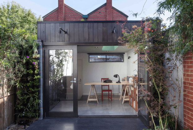 A compact backyard studio or home office opening onto a dark timber deck and enclosed by lush planting. The modern addition has a black vertical timber-clad upper fascia, pale rendered walls, full-height glazed double doors, and a narrow horizontal clerestory window. Inside is a simple workspace with a timber trestle desk, red chair, desk lamp, shelving, and polished concrete flooring. Red brick gabled roofs of the original or neighbouring structure are visible behind it.