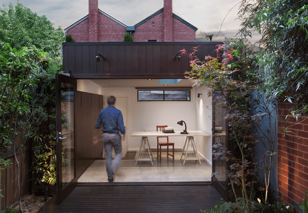 A compact contemporary courtyard extension viewed from a dark timber deck. The open glazed doors reveal a bright kitchen or kitchenette with a pale benchtop, dark timber cabinetry, sink and tap, and a small dining table. Above, a dark-clad upper structure bridges the courtyard, backed by red brick gabled roof forms and chimneys. Dense planted borders frame the space, creating a sheltered indoor-outdoor connection.