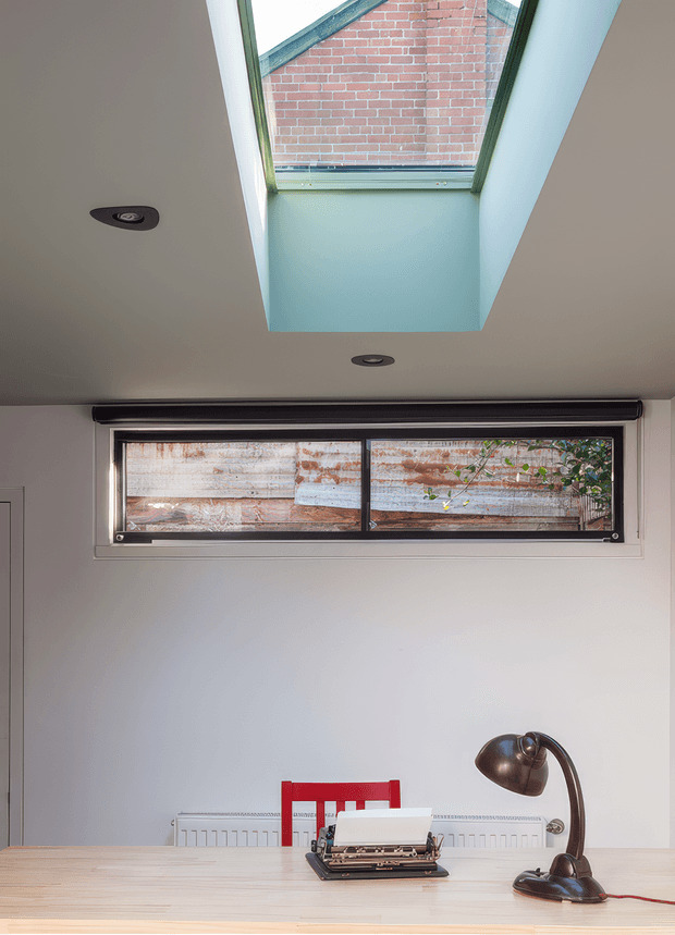 A compact, light-filled home office with a pale timber desk, vintage typewriter, curved dark desk lamp, and red chair. The room has smooth light walls, a narrow horizontal window, recessed downlights, and a prominent roof skylight revealing an adjacent brick wall and greenery outside. The styling combines clean contemporary architecture with vintage-industrial furnishings.