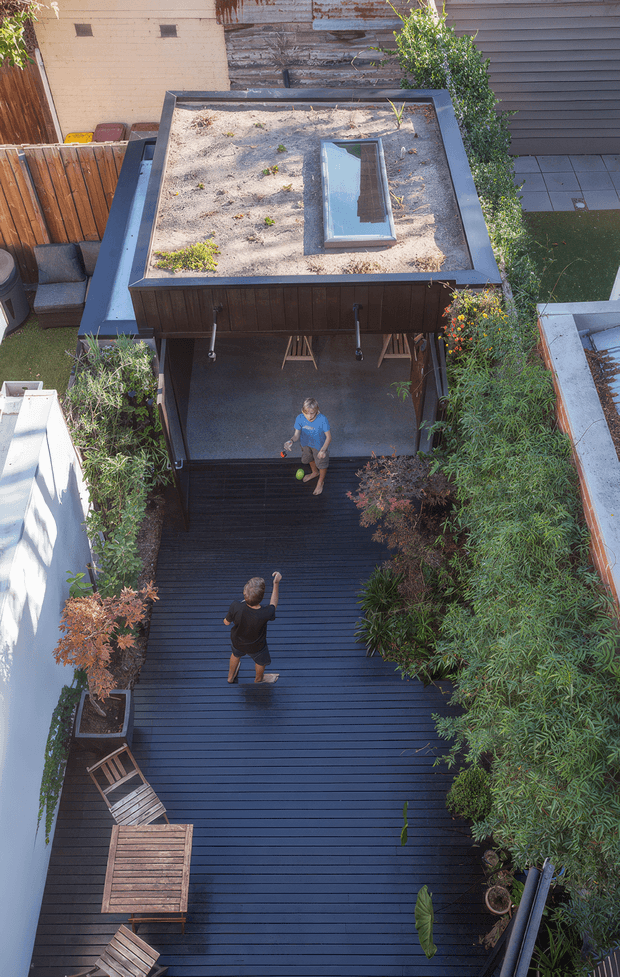 Aerial view of a narrow landscaped courtyard and outdoor entertaining area. A dark timber deck runs between dense planting and rendered boundary walls, leading to a covered pavilion with a planted green roof and a rectangular skylight. The pavilion appears to contain an outdoor seating or dining zone, with steps connecting the upper deck levels. Two people are visible on the deck, along with timber outdoor furniture and large planters. The design has a contemporary urban courtyard character with strong linear decking, integrated greenery, and a green-roofed structure.