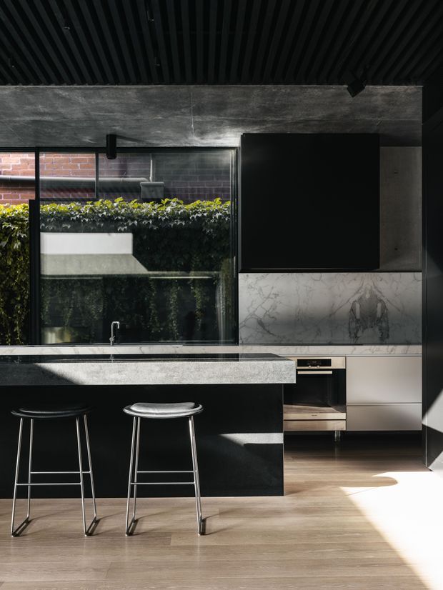 A dark, contemporary kitchen with a large black island and two metal stools, set against full-height glazing overlooking greenery and brickwork. The kitchen features sleek dark cabinetry, a pale veined stone splashback, integrated appliances, timber-look flooring, and a black slatted ceiling. Strong sunlight creates contrasting patches of light across the floor and joinery.