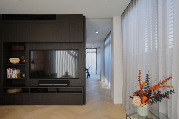 A contemporary interior view centred on a dark timber built-in media wall with television, open display shelving and concealed storage. The space opens into a bright corridor or living area with full-height windows, sheer curtains and light herringbone timber flooring. A glass side table with a vase of orange foliage adds a warm accent.