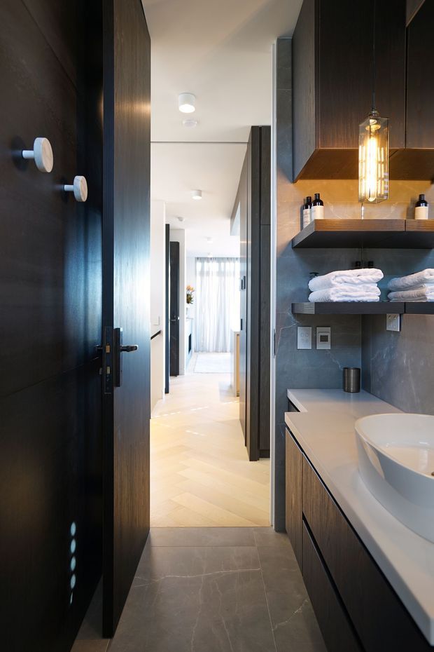 A compact, contemporary bathroom viewed from the doorway, with dark timber cabinetry and door, grey stone-look wall and floor tiles, open timber shelves holding folded white towels, a white countertop basin, and a warm vertical pendant light. The bathroom opens onto a bright hallway with pale herringbone timber flooring.