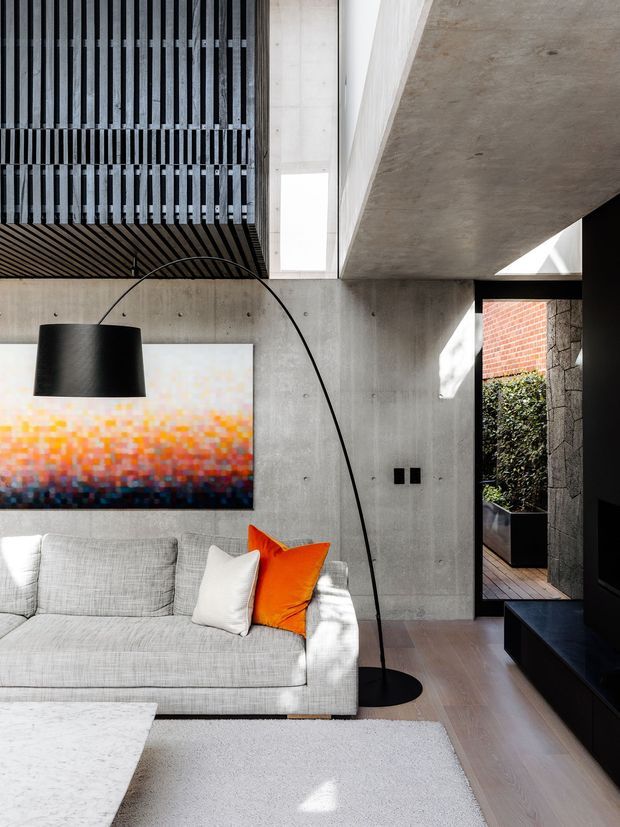 Contemporary living room with a light grey sectional sofa, sculptural black arc floor lamp, colourful abstract artwork, pale timber flooring, and a large light rug. The room features exposed raw concrete walls and ceiling, a dark slatted ceiling section, and a partially visible brick-and-stone courtyard through the opening. Strong natural light creates a refined but slightly industrial atmosphere.