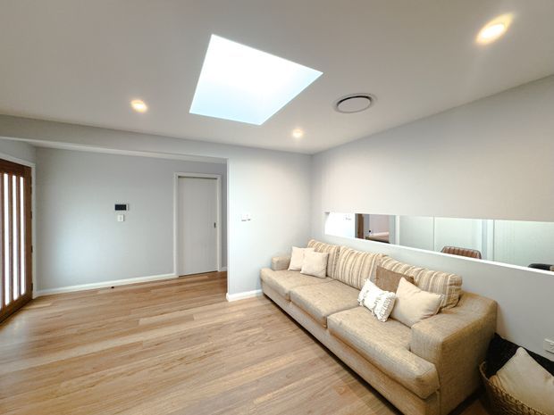 Bright, contemporary living room with light timber flooring, pale grey walls, recessed ceiling lights, and a large rectangular skylight. A substantial beige sectional sofa sits against a half-height mirrored wall, with a doorway and timber-framed glazing at the left.