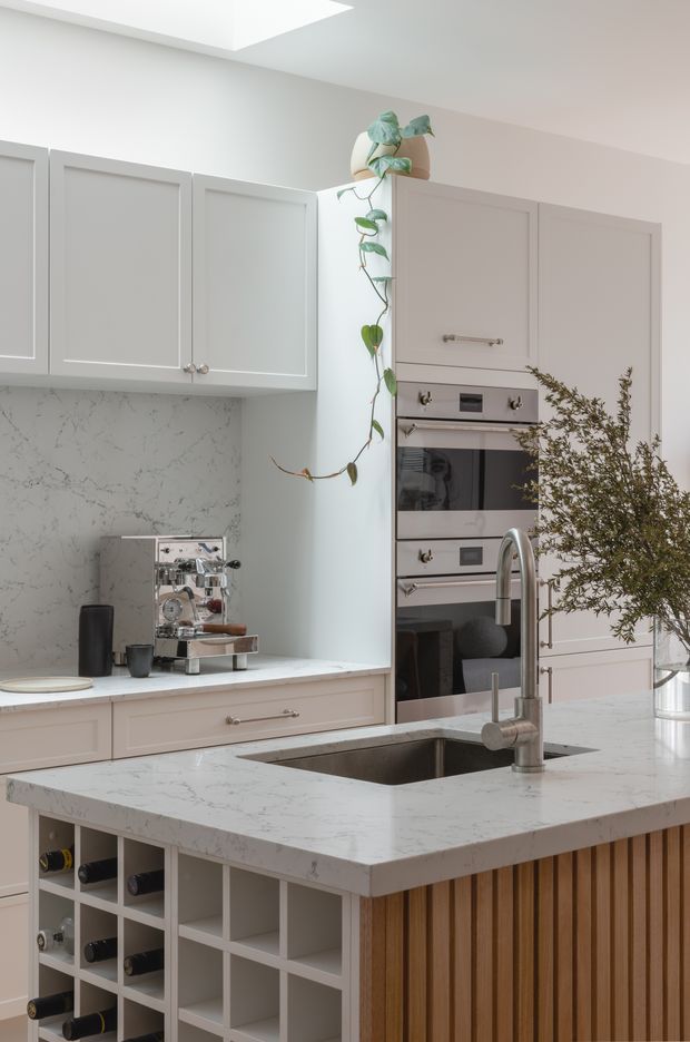A bright, refined kitchen featuring pale grey shaker-style cabinetry, a large marble-look island with integrated sink and brushed metal tapware, stainless steel built-in ovens, and open wine storage. The island has a vertical timber slat detail, while a trailing plant, coffee machine, and branch arrangement add soft natural accents. A skylight or rooflight brings in abundant daylight.