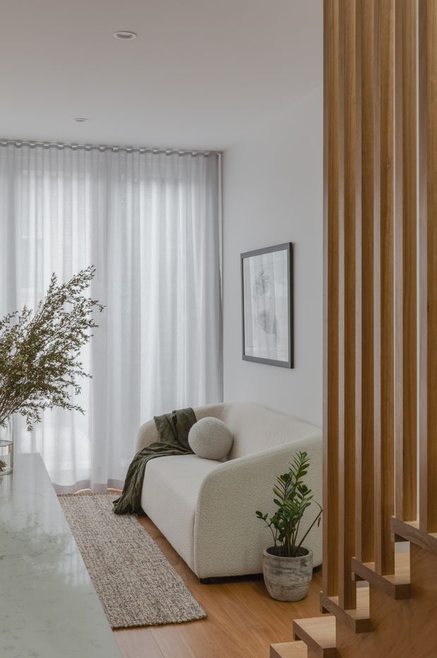 A calm, light-filled living room with a curved cream sofa, round cushion, textured throw, indoor plants, sheer full-height curtains, and a pale area rug over warm timber flooring. A large marble-look surface is visible in the foreground, while a vertical timber slat screen frames an open stairway at the right. The room has white walls, recessed ceiling lights, and understated artwork.