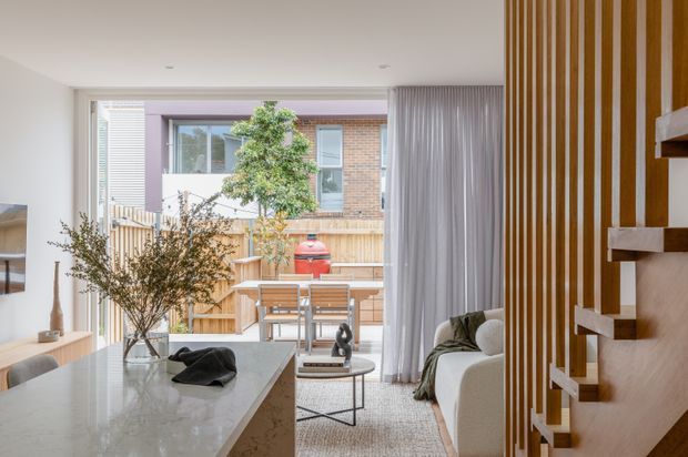 Bright contemporary open-plan living area with a kitchen island in the foreground, a dining setting and lounge beyond, and a compact outdoor courtyard visible through full-height glazing. A sculptural timber slat wall and floating stair treads form a strong feature along the right side. Neutral finishes, sheer curtains, indoor planting, and abundant natural light create an airy, refined interior.