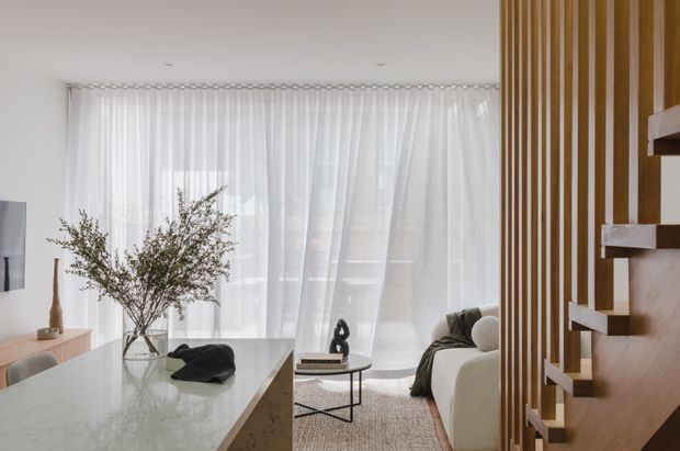 Bright contemporary open-plan interior with a pale stone kitchen island in the foreground, sheer full-height curtains across broad glazing, and a softly furnished living area beyond. A sculptural open-riser timber staircase with vertical slatted screening forms a strong architectural feature on the right. The palette is light and neutral with warm timber accents, a textured rug, and restrained decor.