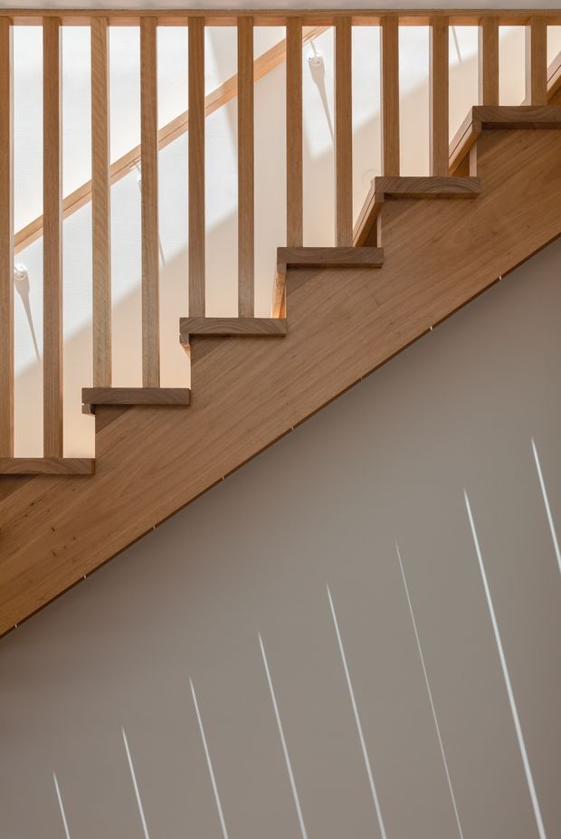 Close-up architectural photograph of an interior timber staircase viewed from the side. A solid warm wood stair stringer and matching treads are paired with evenly spaced vertical timber balusters, creating a clean geometric rhythm. White walls and strong natural light produce diagonal shadows across the lower wall.